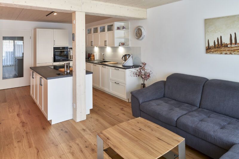 Moderne offene Küche mit weißen Schränken, einer zentralen Kücheninsel und einem grauen Sofa im Wohnbereich, Holzboden und schlichte Dekoration.