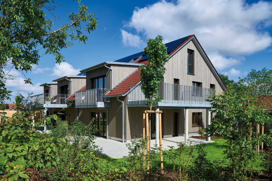 Zweistöckiges Holzhaus mit roten Ziegeldächern und Solarpanelen, umgeben von grüner Landschaft und blühenden Sträuchern, unter blauem Himmel.