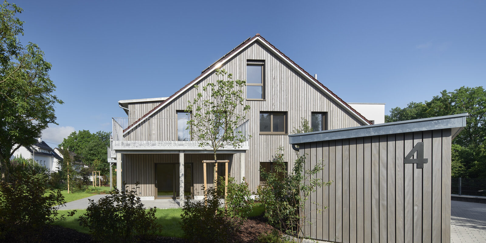 Modernes, zweistöckiges Holzhaus mit großen Fenstern und einem kleinen Schuppen mit der Nummer 4 im Vordergrund, umgeben von gepflegtem Garten und Bäumen unter blauem Himmel.