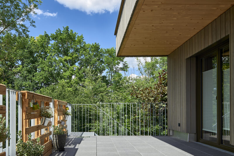 Moderne Terrasse mit grauen Fliesen, umgeben von grüner Natur, mit hölzernem Geländer und Blumentöpfen; blauer Himmel und weiße Wolken im Hintergrund.