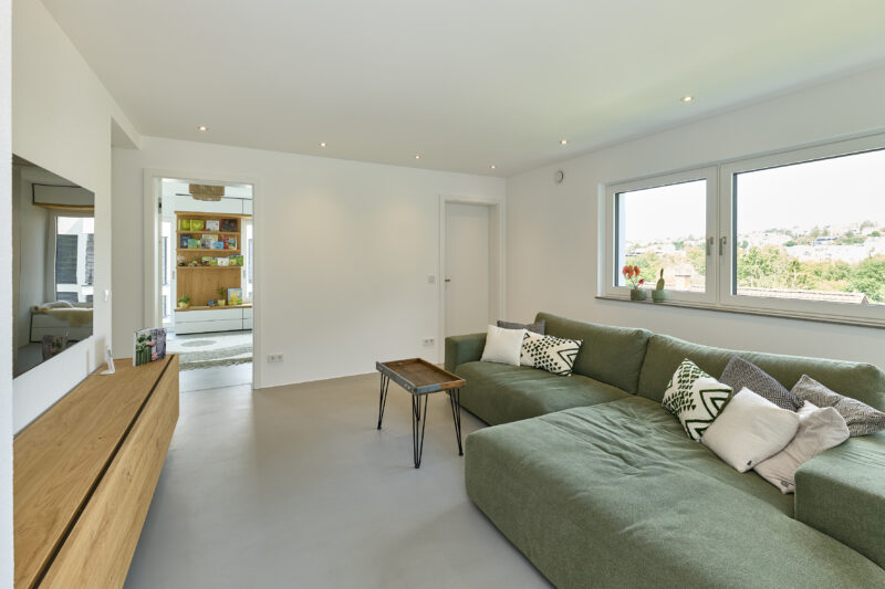 Modernes Wohnzimmer mit grüner Eckcouch, dekorativen Kissen, kleinem Holztisch und einem großen Fenster mit Ausblick auf eine grüne Landschaft.