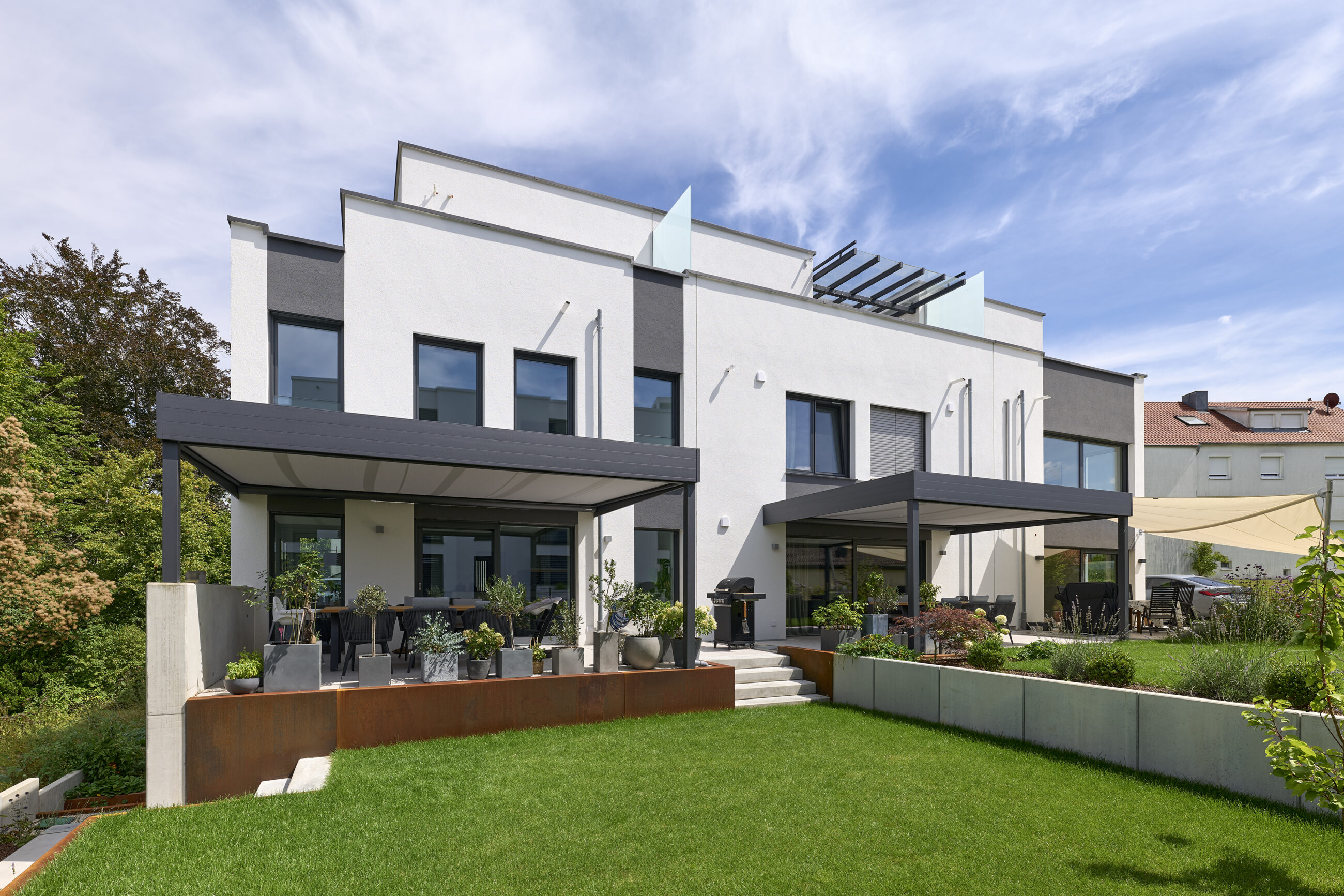 Modernes zweistöckiges Gebäude mit weißer Fassade, großen Fenstern und zwei überdachten Terrassen, umgeben von einem gepflegten Garten.