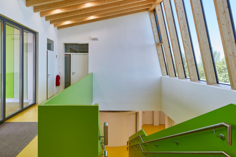 Modernes Treppenhaus mit grünen Geländern, großen Glasfenstern und Holzdecke, das viel natürliches Licht hineinlässt.