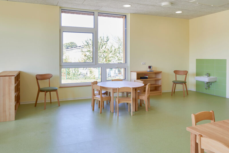 Heller Kindergartenraum mit Holzmöbeln, rundem Tisch und Stühlen vor großen Fenstern, grünem Boden und Waschbecken.