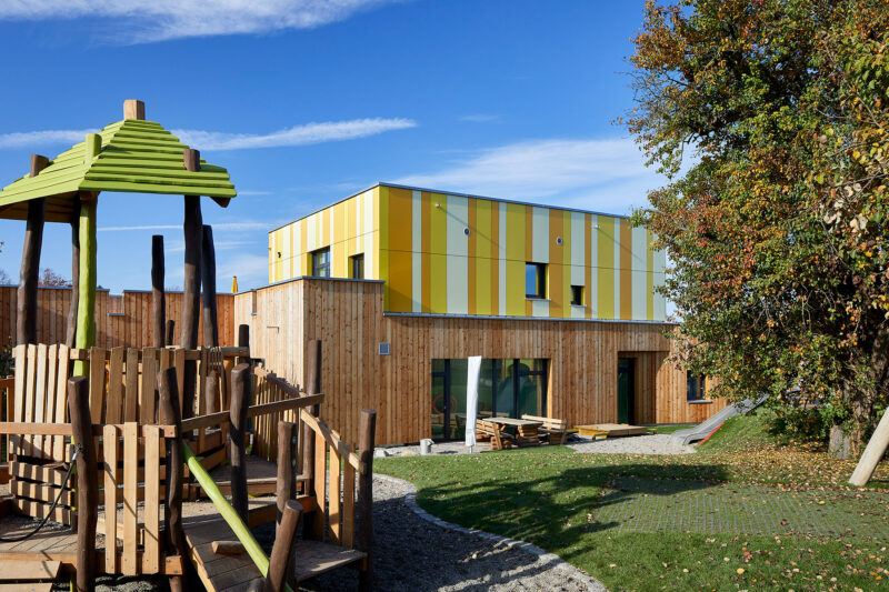 Moderne Kindertagesstätte mit gelb-grüner Fassade und Holzelementen, umgeben von einem Spielplatz mit Holzstrukturen und einem großen Baum im Vordergrund.