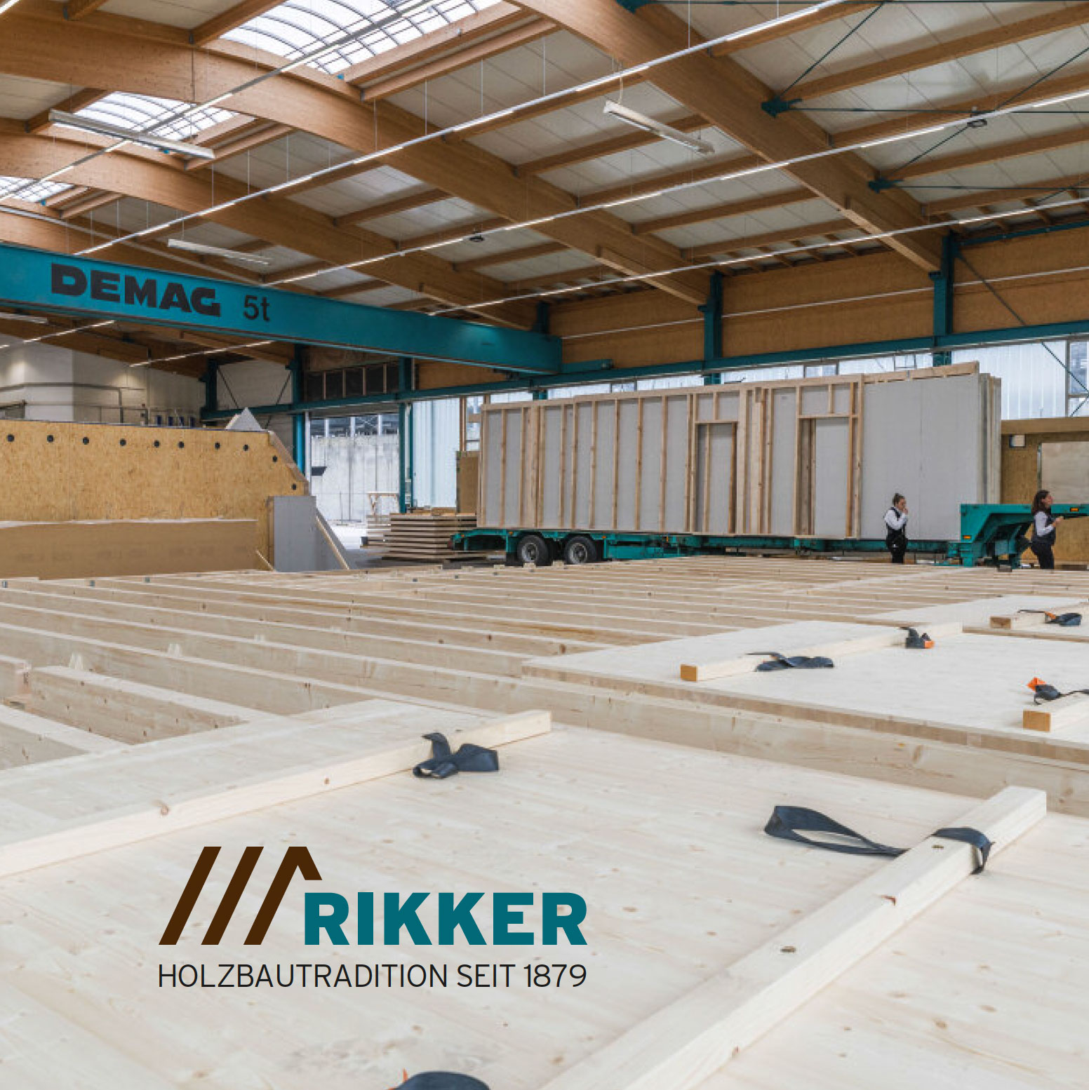 Holzbauhalle der RIKKER Haus GmbH mit Holzrahmenkonstruktionen und einem Kran der Marke DEMAG; im Hintergrund wird eine große Holzwand auf einen Anhänger geladen.