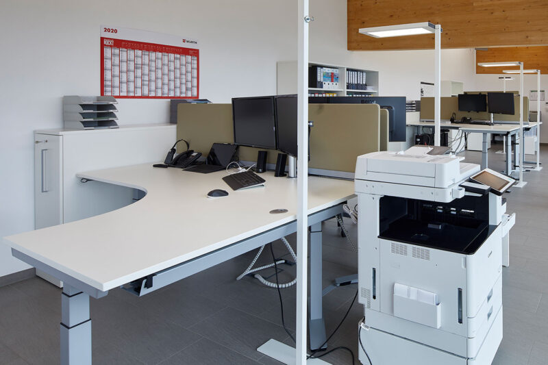 Moderne Büroeinrichtung mit weißen höhenverstellbaren Schreibtischen, mehreren Monitoren und einem multifunktionalen Drucker unter LED-Deckenbeleuchtung.