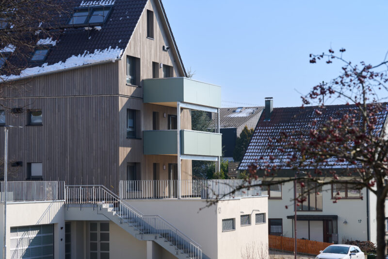Mehrfamilienhaus mit moderner Holzfassade, zwei Balkonen und Außentreppe, im Hintergrund weitere Wohngebäude mit verschneiten Dächern.