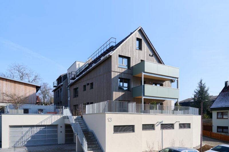 Modernes Mehrfamilienhaus mit Holzfassade, Balkonen und Garage, umgeben von anderen Wohngebäuden, unter blauem Himmel.