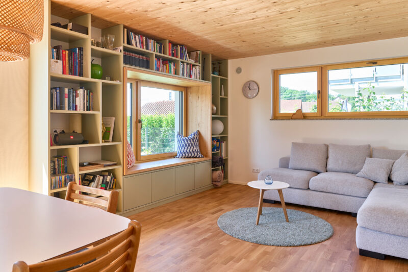 Modernes Wohnzimmer mit hellen Holzmöbeln, einer großen grauen Couch, einem runden Teppich, einem Beistelltisch und raumhohen Bücherregalen neben einem großen Fenster mit Sitzbank.