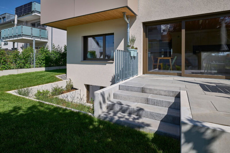 Moderner Außenbereich eines Hauses mit Terrasse, Treppe und gepflegtem Garten; im Hintergrund sind Balkone eines benachbarten Gebäudes zu sehen.