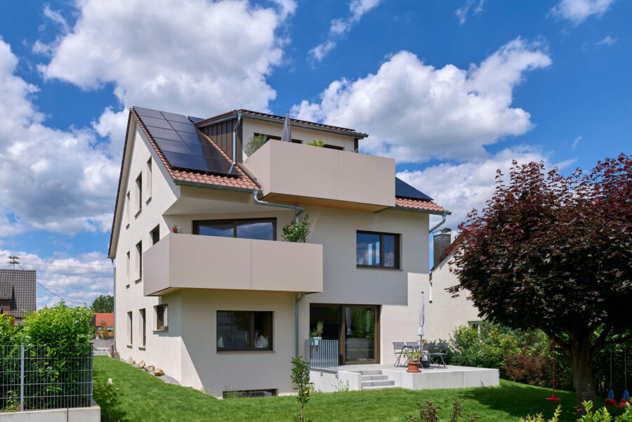 Modernes Mehrfamilienhaus mit Solaranlagen auf dem Dach, einem großen Balkon, gepflegtem Garten und Terrasse, blauer Himmel im Hintergrund.