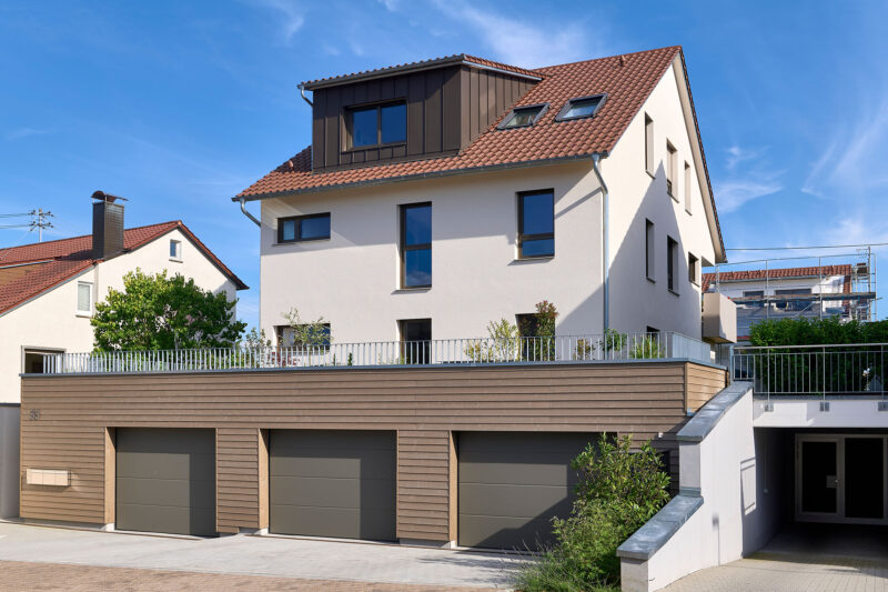 Modernes Einfamilienhaus mit rotem Satteldach und weißer Fassade, ergänzt durch eine dreifach integrierte Garage mit Holzverkleidung und angrenzendem Gartenbereich.