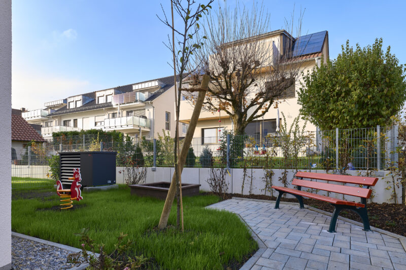 Garten eines Wohnkomplexes mit einer roten Bank, einem Wipptier und einem gepflasterten Weg, im Hintergrund moderne Gebäude mit Solarpanelen.