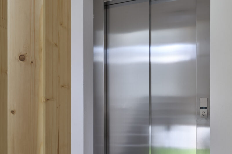 Silberner Aufzug mit geschlossenen Türen in einem modernen Flur mit Holzwand und gepunkteter Decke.